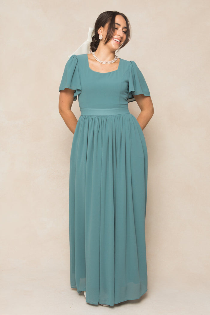 A woman wearing the Ivy City Co Abbie Dress in Seaglass Chiffon—a floor-length gown in a teal hue featuring flutter sleeves—stands smiling against a beige background, hands tucked into her dress pockets. She accessorizes with pearl jewelry and a white ribbon in her hair, creating an elegant look perfect for bridesmaids.