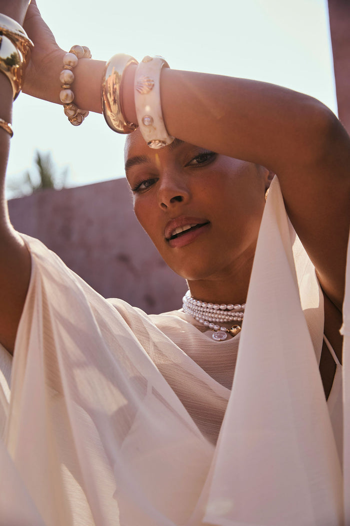 Layered pearl and gold choker worn with an ivory chiffon cape dress in sunlight.