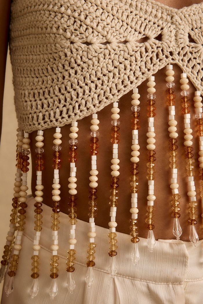 Close-up of cream crochet festival top with amber beaded fringe hem over white pants.
