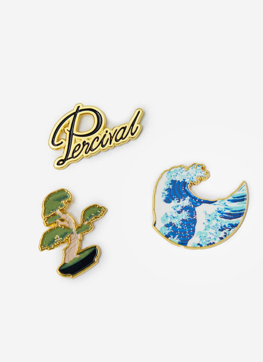 A collection of three gold-trimmed metal pin badges by Percival, showing the brand logo, a bonsai tree and a Japanese wave.