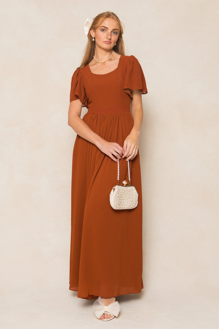 A woman in the Abbie Dress in Amber Chiffon by Ivy City Co, a short-sleeve maxi, holds a small textured cream purse. She pairs it with white sandals and a white hair bow, standing against a neutral background.