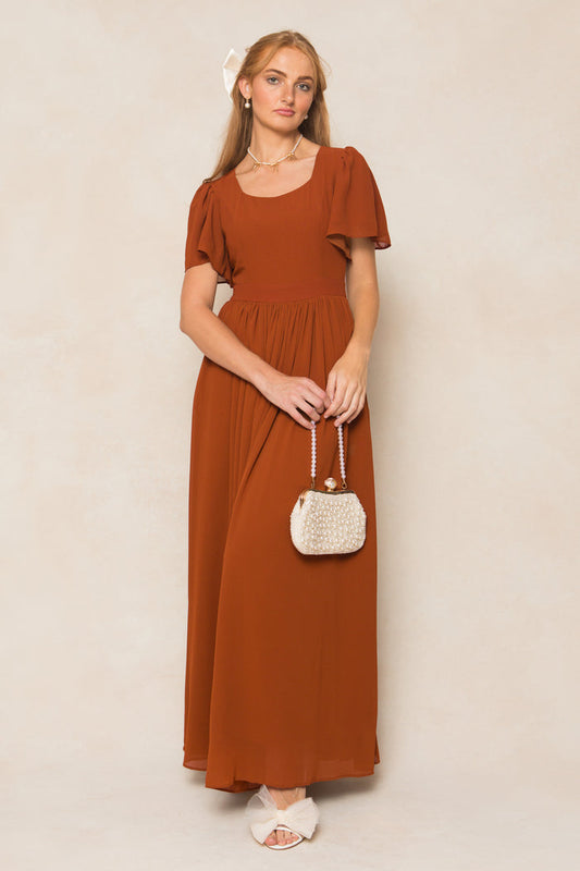 A woman in the Abbie Dress in Amber Chiffon by Ivy City Co, a short-sleeve maxi, holds a small textured cream purse. She pairs it with white sandals and a white hair bow, standing against a neutral background.