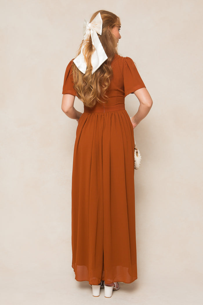 A woman with long hair and a white bow stands with her back to the camera in the Ivy City Co Abbie Dress in Amber Chiffon, a short-sleeve maxi dress, paired with a small white purse and white platform heels.