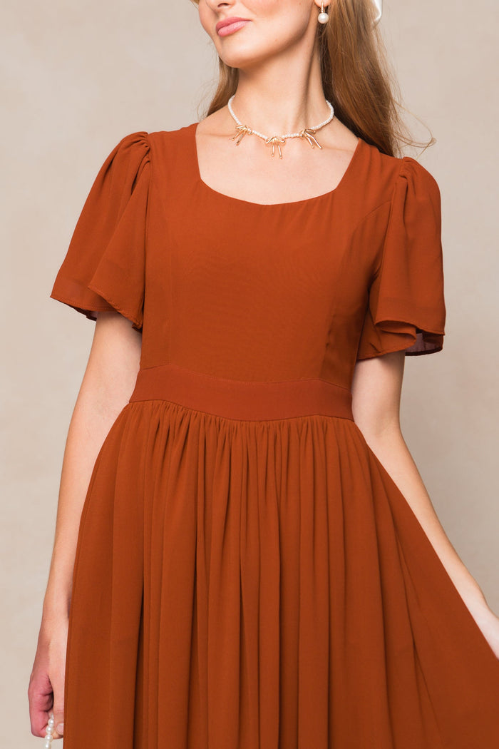 A woman poses against a neutral background wearing the Ivy City Co Abbie Dress in Amber Chiffon—a rust-colored, short-sleeve dress with a square neckline and gathered skirt reminiscent of a classic chiffon bridesmaid dress—paired with a pearl necklace featuring a bow detail.