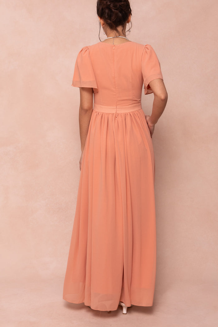 A woman stands with her back to the camera in the Ivy City Co Abbie Dress in Apricot Crush, a short-sleeved maxi with a gathered skirt and fitted waist, ideal for bridesmaids, set against a soft pink background.