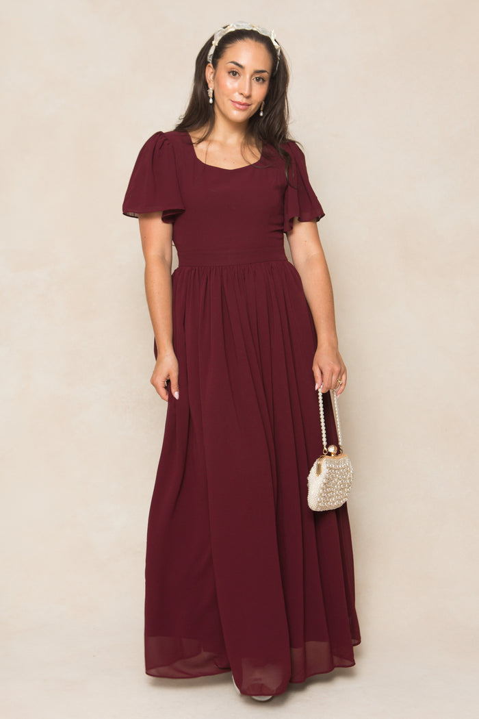A woman in the Ivy City Co Abbie Dress in Wine Chiffon, a short-sleeve, floor-length bridesmaid dress, stands against a light background holding a beaded purse and wearing a headband with her hair down and a neutral expression.