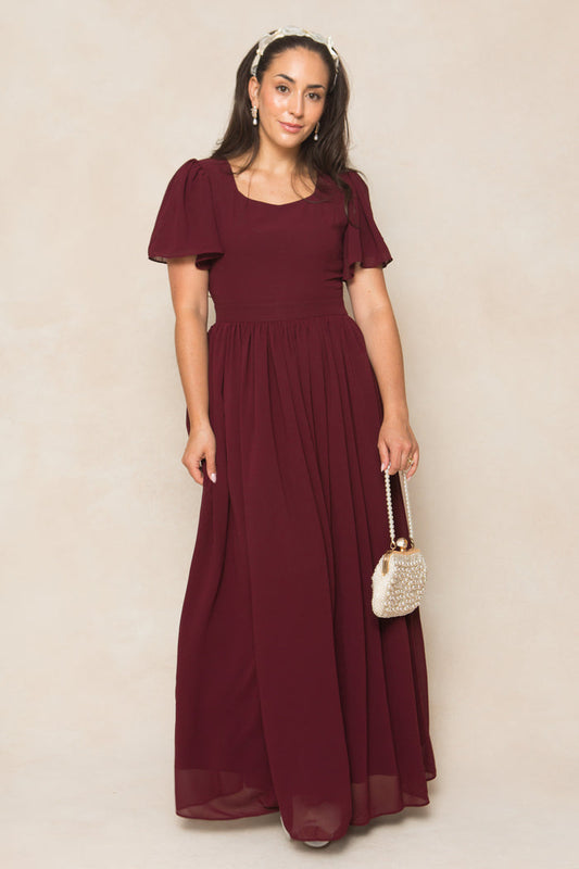 A woman in the Ivy City Co Abbie Dress in Wine Chiffon, a short-sleeve, floor-length bridesmaid dress, stands against a light background holding a beaded purse and wearing a headband with her hair down and a neutral expression.