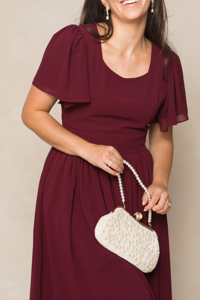 A woman smiles in the Ivy City Co Abbie Dress in Wine Chiffon, holding a small white beaded purse. She wears pearl earrings and her brown hair is down. The image captures her from the shoulders to the waist.