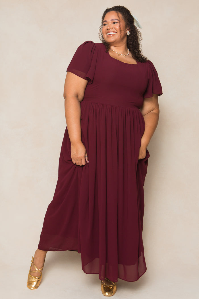 A woman with curly hair smiles to the side, standing against a light beige background in the Ivy City Co Abbie Dress in Wine Chiffon—flowing, short-sleeved, with one hand in her pocket and gold shoes.