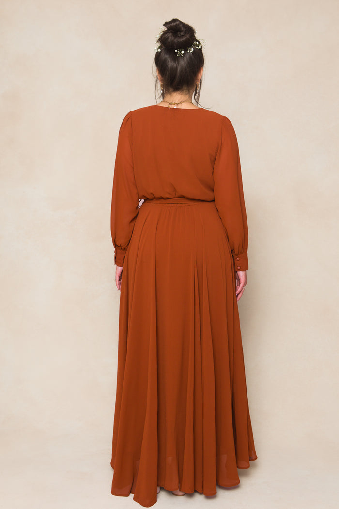 A woman faces a beige wall with her back to the camera, wearing the Andie Dress in Amber Chiffon by Ivy City Co. Her hair is styled in a bun with small white flowers, creating a perfect look for fall bridesmaids.