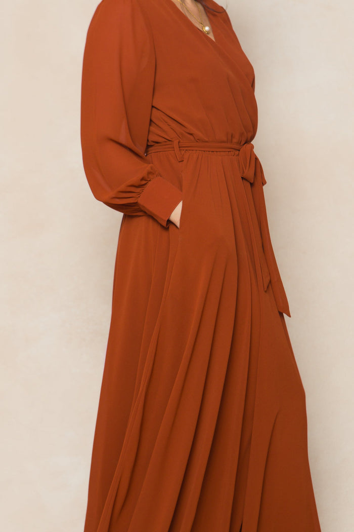 A woman models the Andie Dress in Amber Chiffon by Ivy City Co, featuring long sleeves, a soft and flowy rust-colored chiffon fabric, and a matching belt tied at the waist. With one hand in the pocket and set against a light, neutral background, this women’s maxi dress blends comfort and elegance—ideal for fall bridesmaids or as an elevated everyday look.