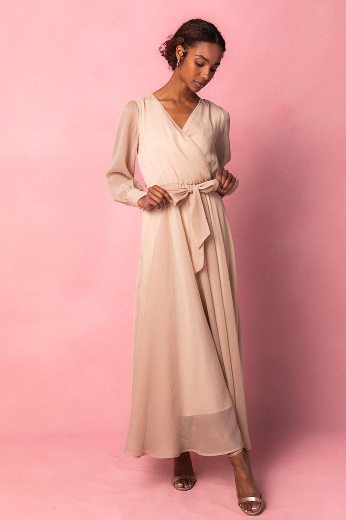 A woman wearing the Ivy City Co Andie Dress in Champagne—a long, light beige maxi dress with a tied waist and long sleeves—stands against a pink background, looking down as she adjusts the dress with one hand, paired with open-toe heels.