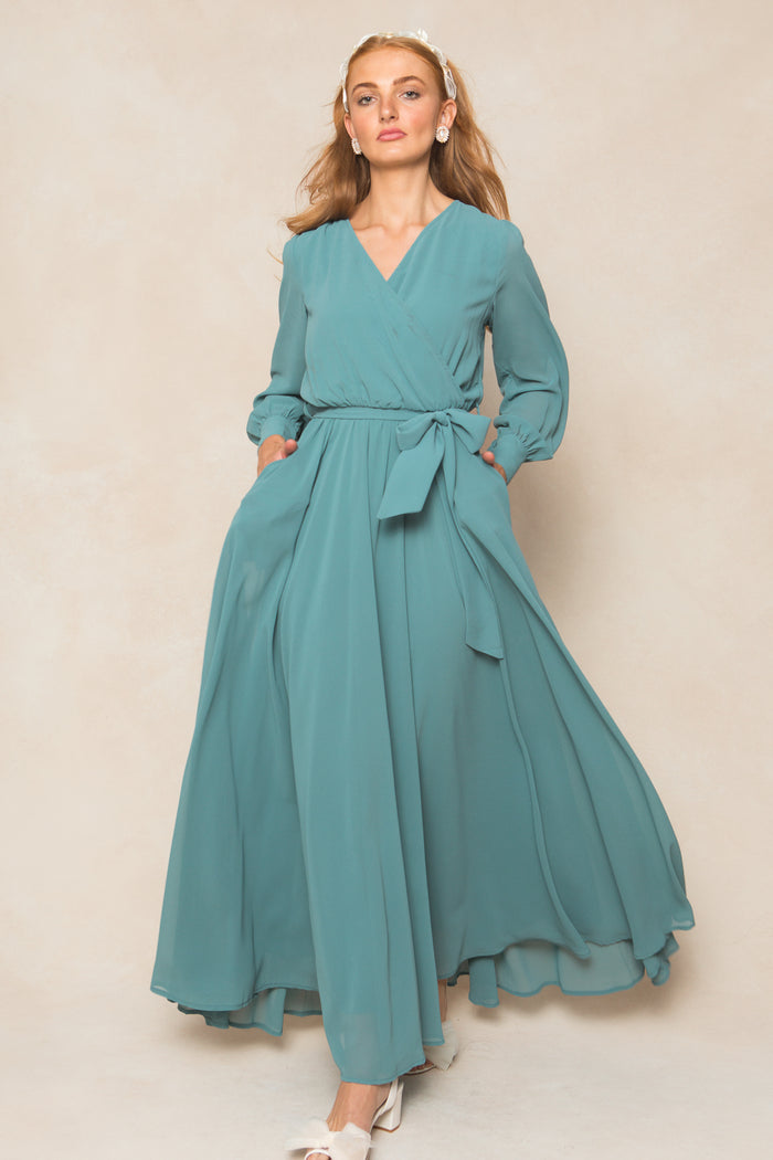 A woman in the Ivy City Co Andie Dress in Seaglass Chiffon, featuring long sleeves and a waist tie, poses against a neutral background. She has long light brown hair, pearl earrings, and white shoes.