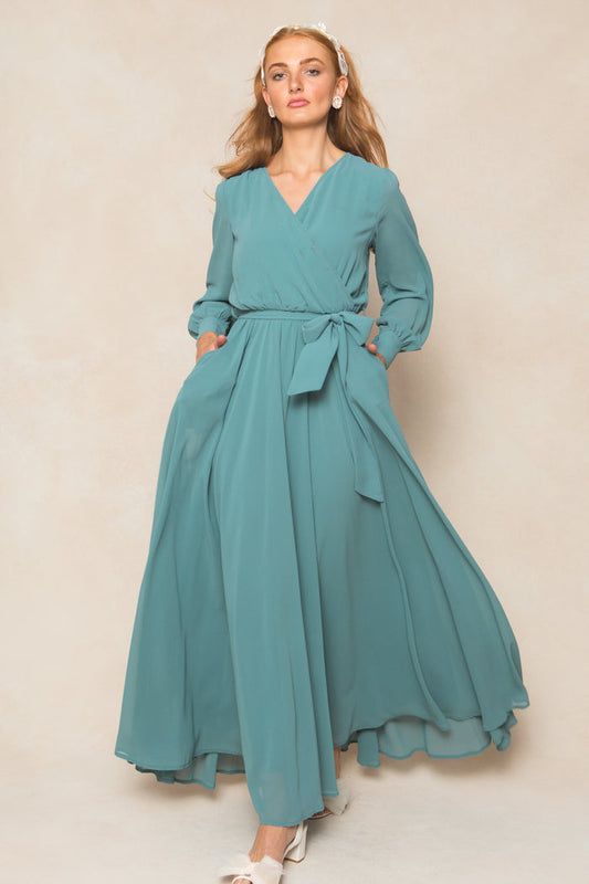 A woman in the Ivy City Co Andie Dress in Seaglass Chiffon, featuring long sleeves and a waist tie, poses against a neutral background. She has long light brown hair, pearl earrings, and white shoes.