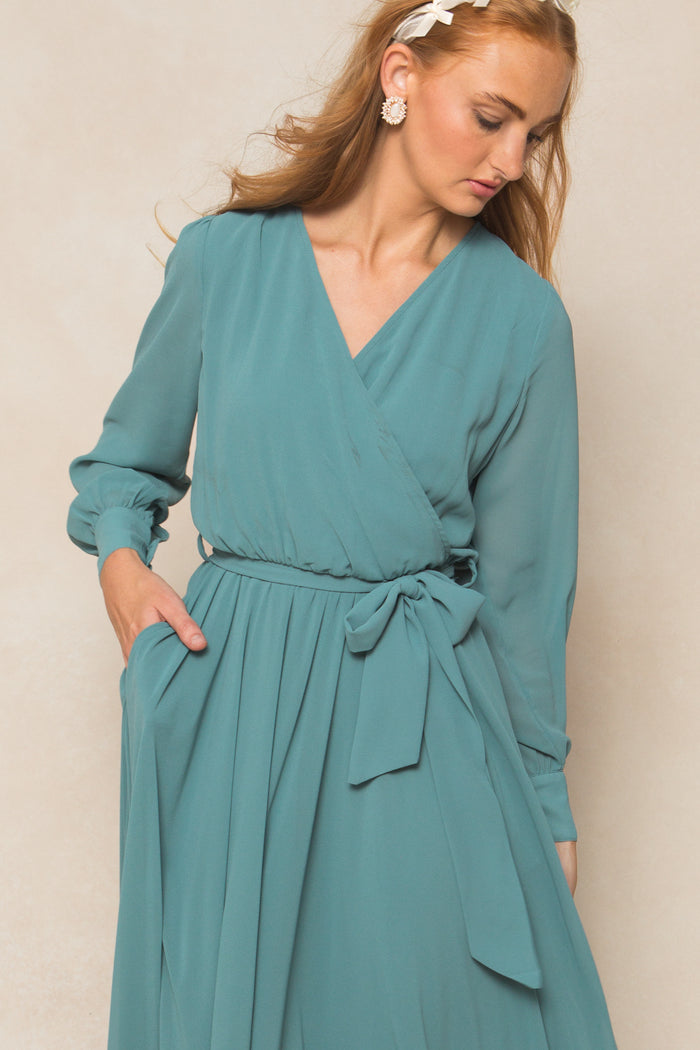 A woman stands against a beige background, looking down with one hand in her pocket, wearing Ivy City Co's Andie Dress in Seaglass Chiffon. The dress features long sleeves, a tied waist bow, and is paired with floral earrings and wavy hair.