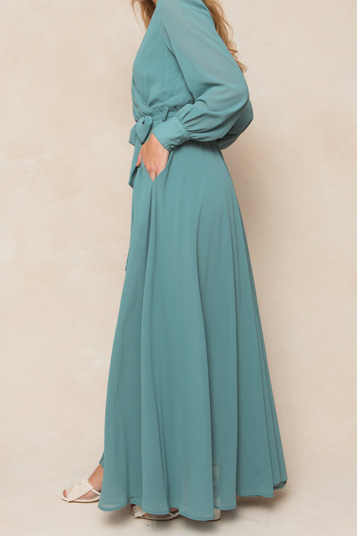 A woman stands sideways in the Ivy City Co Andie Dress in Seaglass Chiffon—an elegant long-sleeve dress with a belted waist and pockets, paired with white open-toe shoes, perfect for fall bridesmaids against a neutral backdrop.