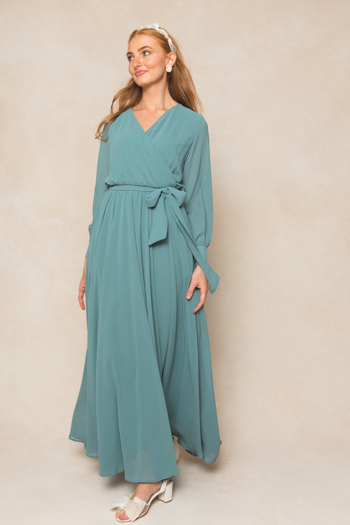 A woman in the Ivy City Co Andie Dress in Seaglass Chiffon, featuring long sleeves and a waist tie, stands against a beige background. She wears white open-toed heels and smiles gently, her long wavy hair falling over her shoulders.