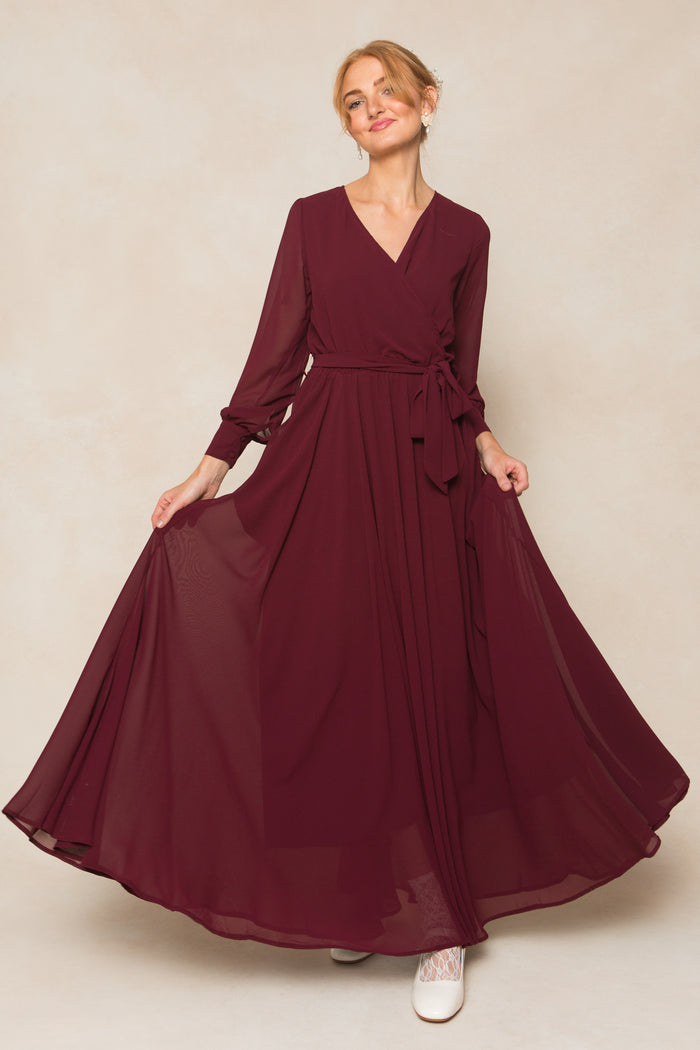 A woman models the Ivy City Co Andie Dress in Wine Chiffon—a long-sleeved maxi with a wrap-style bodice and tied waist—standing against a neutral background, gently smiling while holding out the flowing skirt.