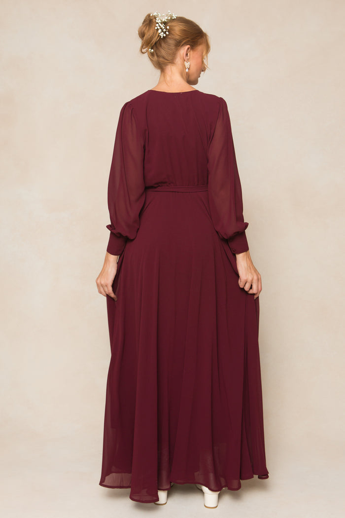 A woman faces away, wearing the Ivy City Co Andie Dress in Wine Chiffon—a long-sleeved, floor-length gown with a flowing skirt. Her light updo is adorned with pearls, paired with light-colored shoes.
