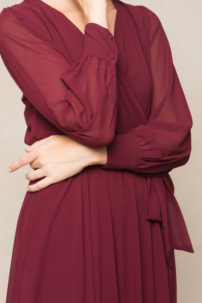 A person wearing the Ivy City Co Andie Dress in Wine Chiffon crosses one arm over their body and touches their chin with the other hand. Their face is partially out of frame.