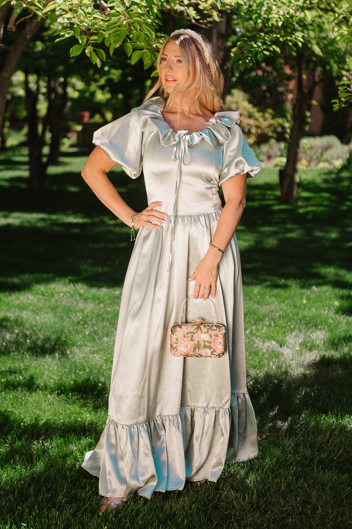 A woman stands on grass under trees, wearing the Ivy City Co Billie Dress in Sage Satin with puff sleeves and ruffles. She holds a small floral purse, looking to the side as sunlight bathes her.