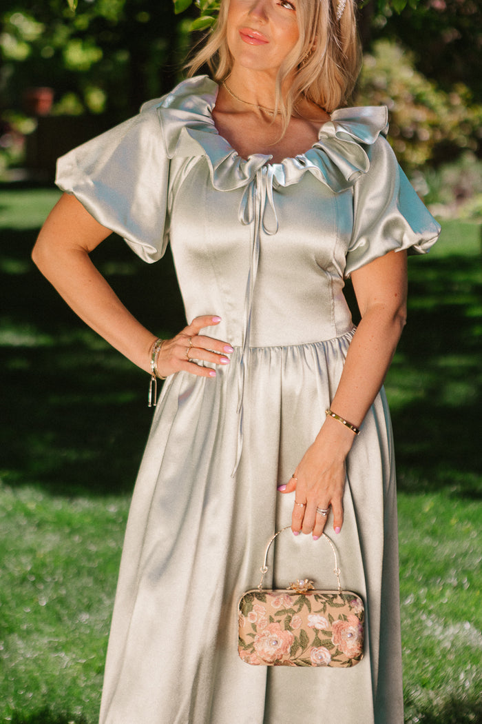 Outdoors on grass, a woman wears the Ivy City Co Billie Dress in Sage Satin with a ruffled neckline and puff sleeves. She holds a small floral-patterned handbag, her nails painted light pink, with sunlight and trees in the background.