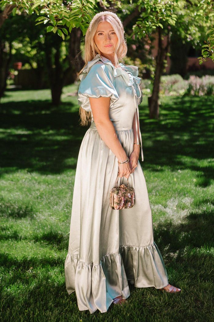 A woman stands on grass in a sunlit garden, wearing the Billie Dress in Sage Satin by Ivy City Co and holding a small floral handbag, with trees and greenery in the background.