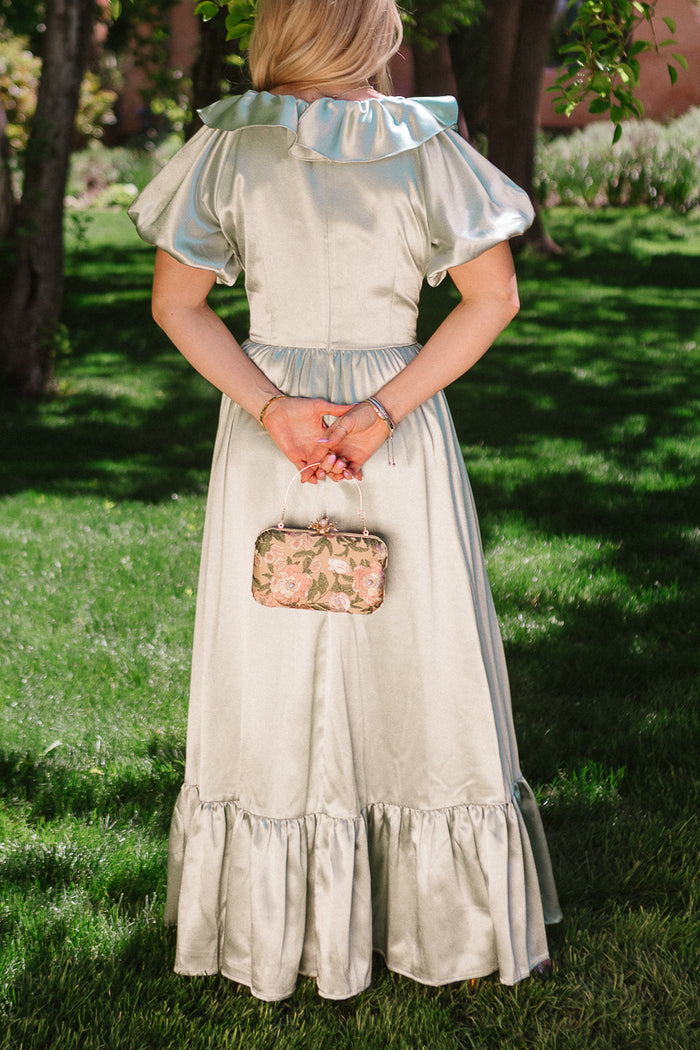 A woman stands on grass in the Ivy City Co Billie Dress in Sage Satin, featuring puffed sleeves and a ruffled hem. Facing away with a floral handbag behind her, she’s surrounded by trees and sunlight for a vintage-inspired look.