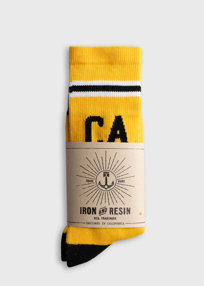 Yellow and black socks with "CA" text, wrapped in Iron and Resin packaging, showcasing a sunburst logo. Designed in California.