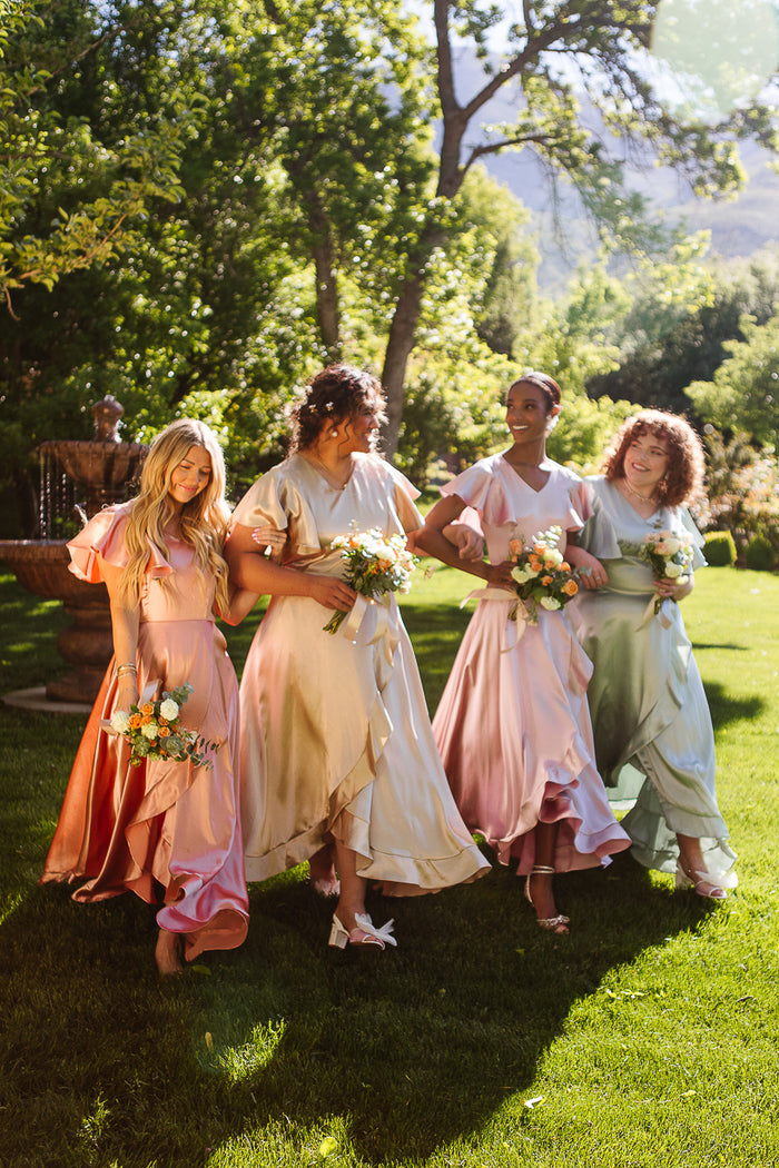 Four women wearing Ivy City Co's Callie Dress in Apricot Crush Satin hold bouquets and walk together on a sunlit lawn, surrounded by greenery, enjoying the outdoor celebration as part of a joyful bridal party.