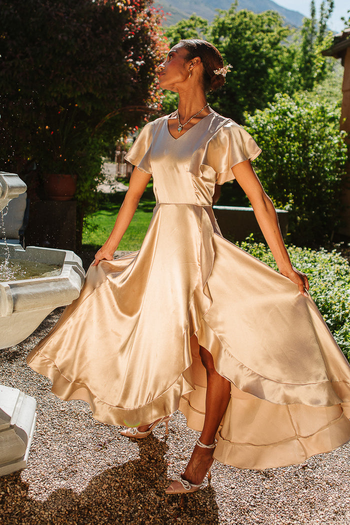 A woman poses gracefully outdoors by a stone fountain, wearing the Ivy City Co Callie Dress in Champagne Satin, its shimmering fabric highlighted by sunlight and lush greenery.