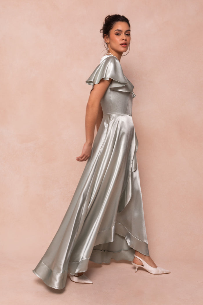 A woman stands sideways in Ivy City Co’s Callie Dress in Sage Satin—shiny, floor-length, with short ruffled sleeves—paired with white pointed-toe heels against a soft beige background.