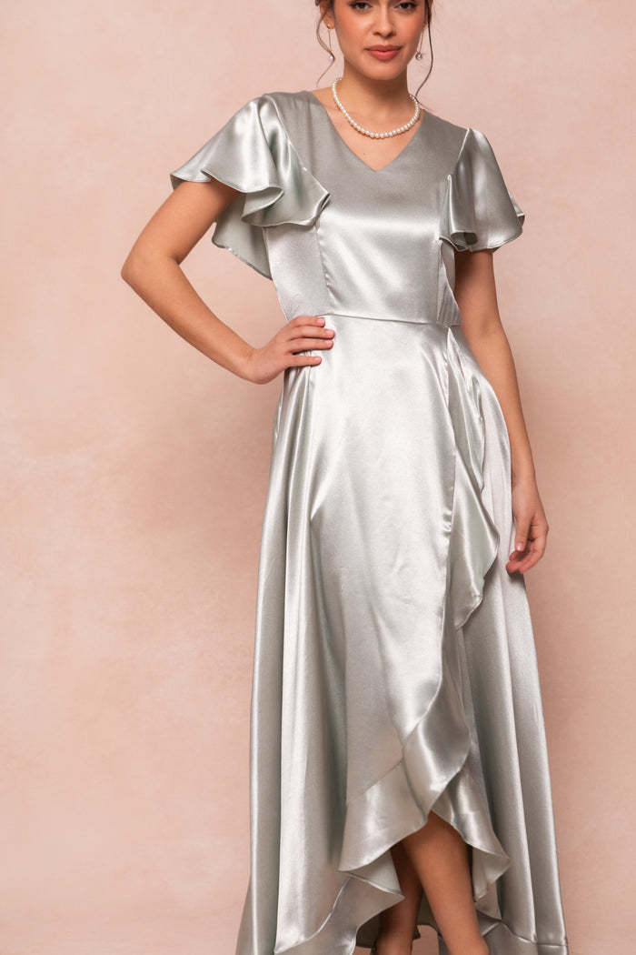 A woman in the Callie Dress in Sage Satin by Ivy City Co, featuring flutter sleeves and a ruffled hem, accessorized with a pearl necklace, stands against a soft beige background.