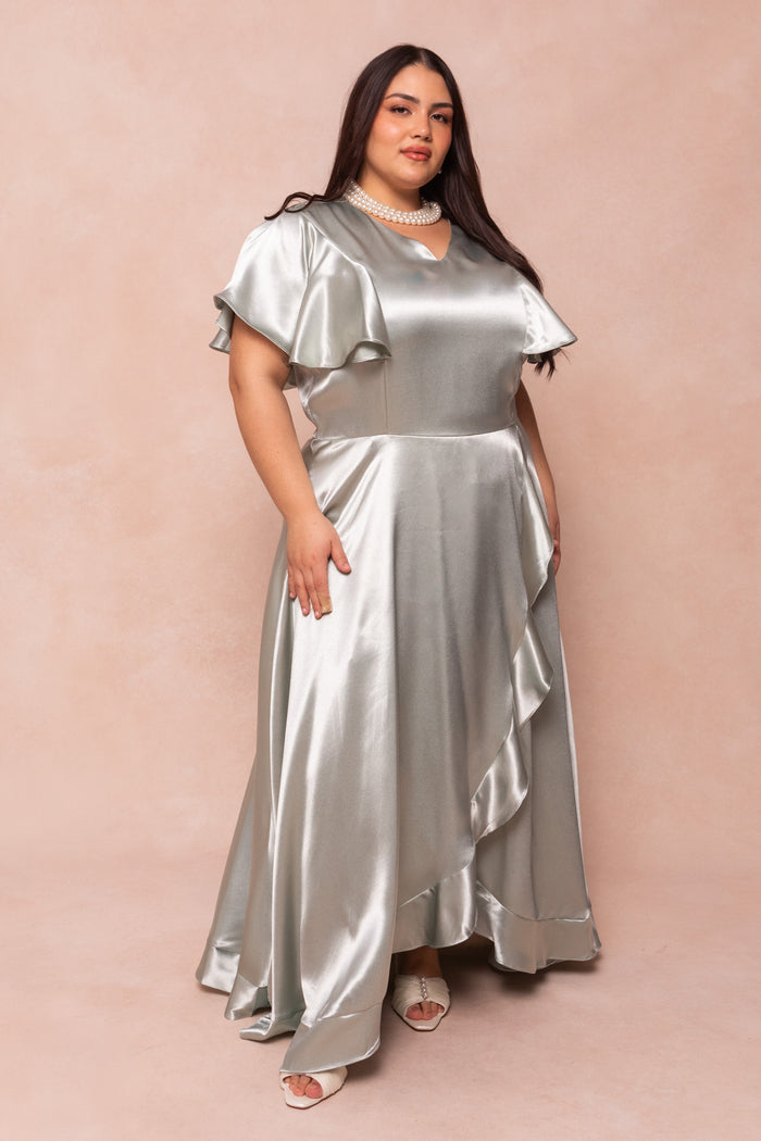 A woman models the Ivy City Co Callie Dress in Sage Satin, featuring short flutter sleeves and a ruffled skirt, paired with white open-toe heels and a pearl necklace against a soft beige background.
