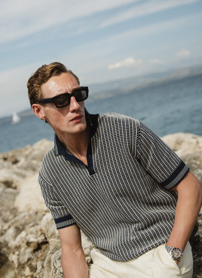 Percival Claude Knitted Polo in navy two-tone textured cotton, worn by a model in a scenic coastal setting.