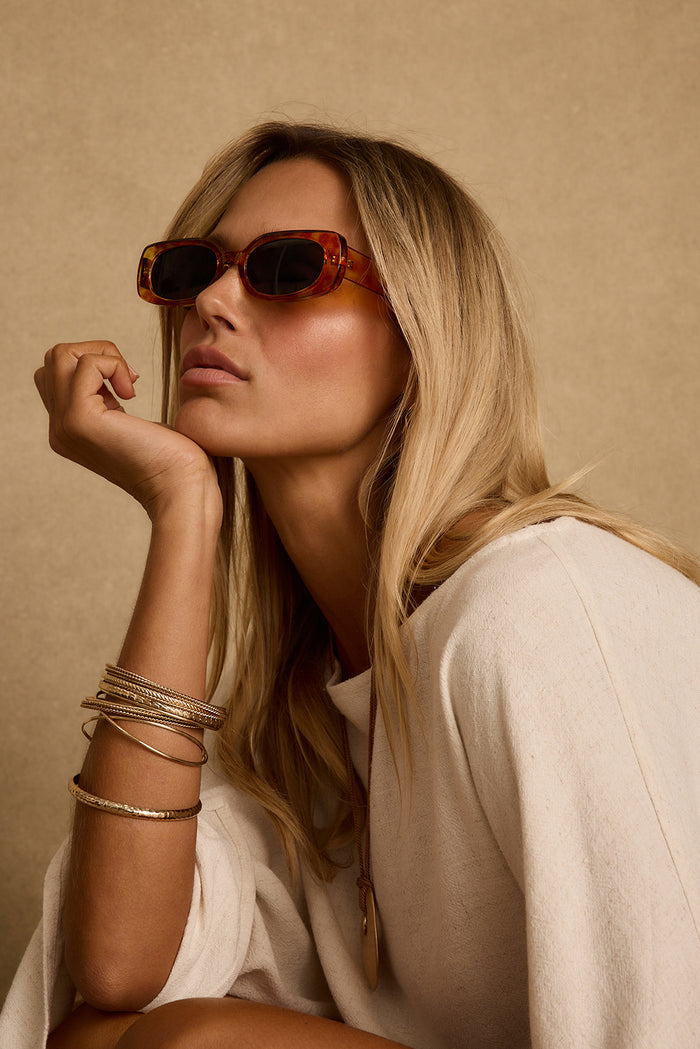 Model wearing rectangular tortoise sunglasses with gold bangles and a cream linen top.