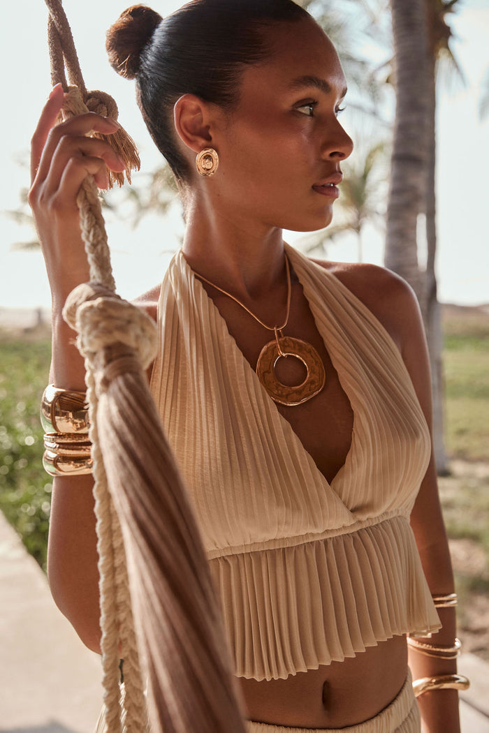 Cream plisse halter crop top styled with gold pendant necklace and cuffs by palm trees.