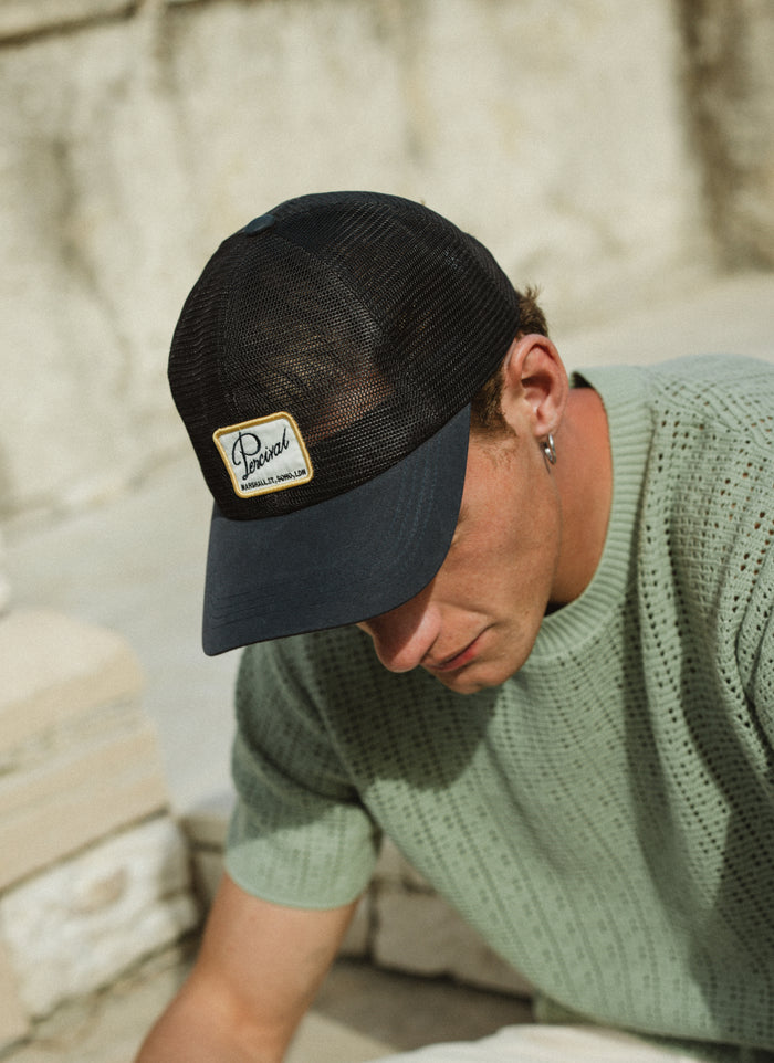 Model wearing the navy Percival Damien Mesh Trucker Cap with a breathable mesh back and a signature woven patch.