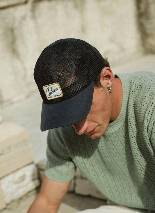 Model wearing the navy Percival Damien Mesh Trucker Cap with a breathable mesh back and a signature woven patch.