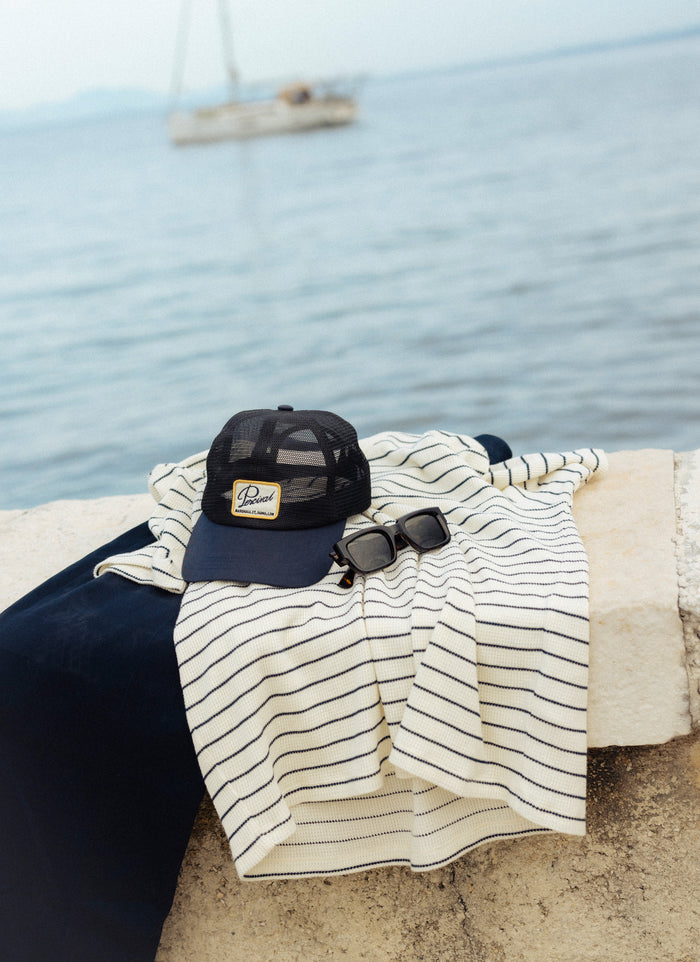 Navy Percival Damien Mesh Trucker Cap with a logo patch, styled with a striped shirt and sunglasses by the coast.