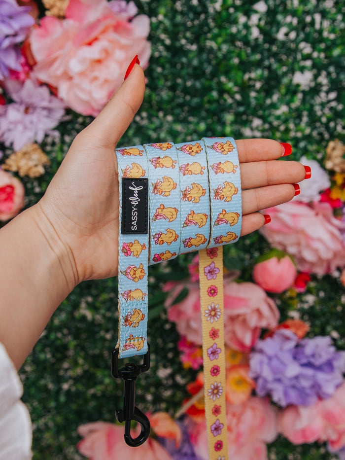 A hand with red-painted nails holds the Sassy Woof "Dog Leash - Good Duck, Babe!" featuring cartoon dogs and flowers on a blue neoprene handle. Blurred colorful flowers and greenery fill the background.