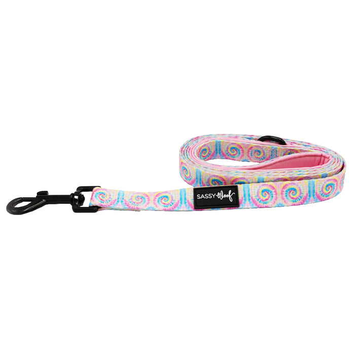 The Sassy Woof Dog Leash - Good Vibes features a pastel swirl tie-dye design, padded handle for comfort, black metal clip, and a Sassy Woof label near the clasp. The leash is coiled to showcase its vibrant style.
