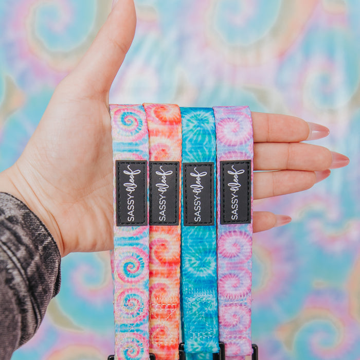 A hand holds the Sassy Woof Dog Leash - Summer Sorbet, a colorful tie-dye leash with a black Sassy Woof label, displayed in front of a pastel swirl-patterned background.