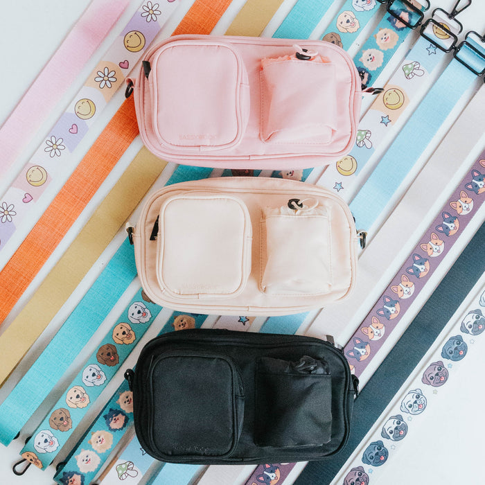 The Sassy Woof Dog Parent Bag + Strap comes in pink, beige, and black with two front pockets and colorful interchangeable straps featuring cute flower, smiley face, and animal designs—perfect as a dog parent bag or treat pouch.