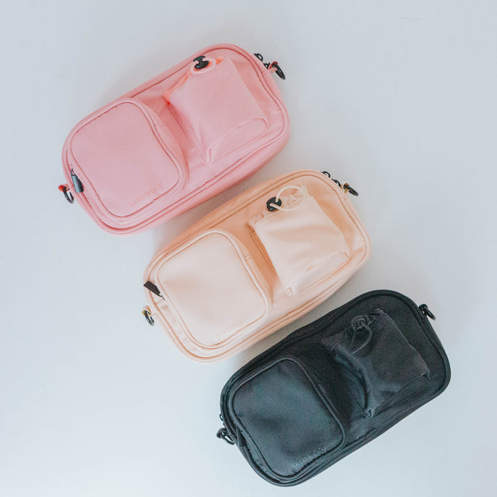 Three Sassy Woof Dog Parent Bags + Straps in pink, beige, and black are arranged on a white surface. Each bag has zipper closure, two front pockets, a small loop on one corner, and interchangeable straps.
