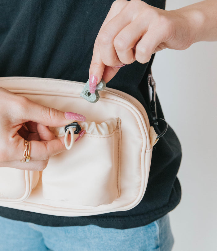 A person with pink glittery nails places a green key-shaped item into the front pocket of the Sassy Woof Dog Parent Bag + Strap, featuring interchangeable straps, while wearing a black shirt and blue jeans.