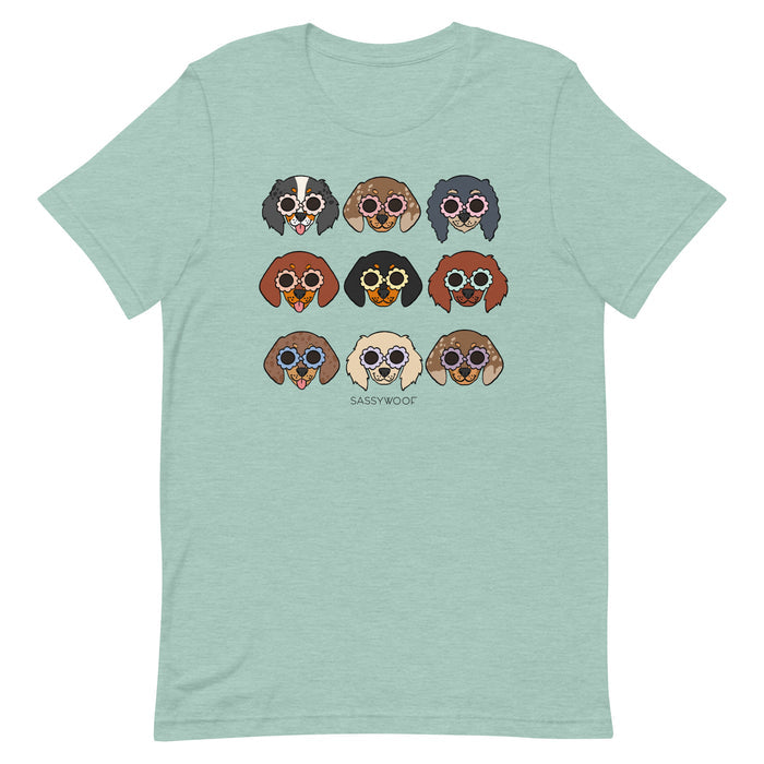 The Doxie Tee by Sassy Woof is a light green unisex 100% cotton t-shirt, featuring nine cartoon dog faces with sunglasses in three rows and "SASSY WOOF" printed underneath. Each pup showcases a unique fur color and pattern.
