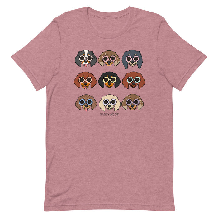 The Doxie Tee by Sassy Woof is a mauve unisex shirt with a grid of nine illustrated dogs in sunglasses, each with unique fur colors and styles. Made from 100% cotton, it features "SASSY WOOF" printed below the dogs.