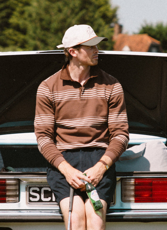 Percival Editor espresso striped rugby polo and cream Spencer cap with rope detail, worn by a man on a vintage car boot.