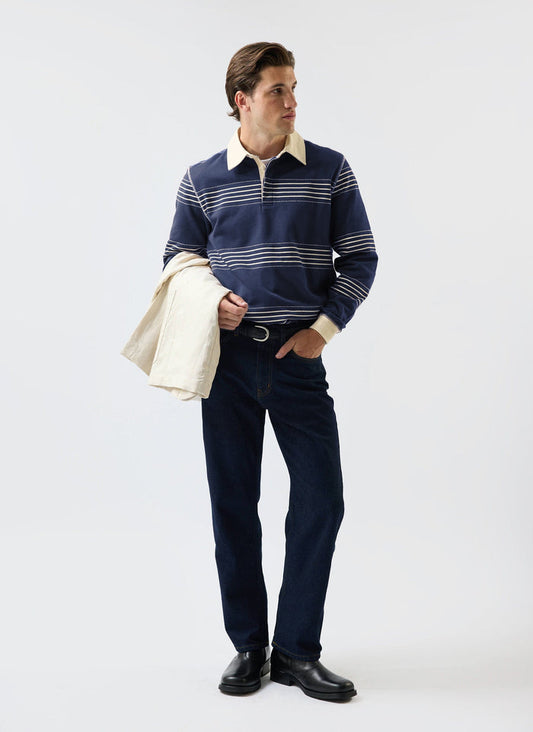 A male model wears the Percival Editor Striped Rugby Polo in navy, styled with dark jeans and a cream jacket over his arm.
